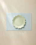 A light green, wavy-edged ceramic bowl sits centered on the Geometry Chambray Light Blue placemat, which rests on a beige tablecloth under soft, natural light.