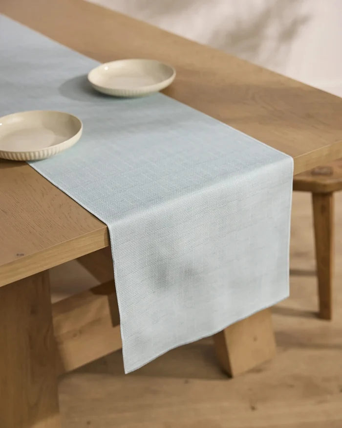 A Chambray Light Blue table runner by Geometry drapes over a wooden dining table topped with two cream-colored bowls. The setting is minimal, complemented by light wood flooring.