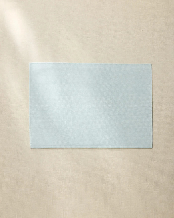 A Geometry Chambray Light Blue rectangular placemat is laid flat on a beige surface, illuminated softly by natural light from the left.