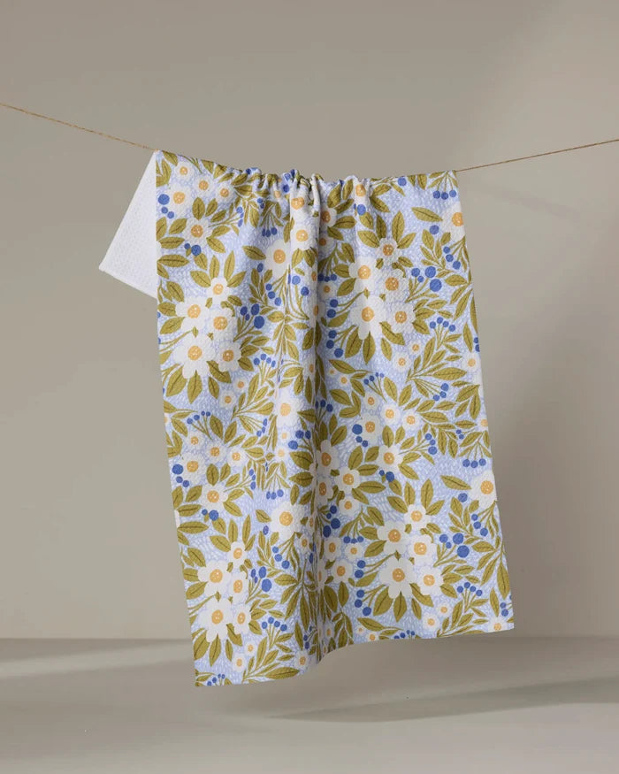 The "Chamomile And Blue" tea towel by Lisa Nelson features a floral pattern in white, green, and blue, crafted from recycled materials and displayed on a clothesline against a neutral background.