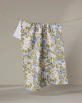 The "Chamomile And Blue" tea towel by Lisa Nelson features a floral pattern in white, green, and blue, crafted from recycled materials and displayed on a clothesline against a neutral background.