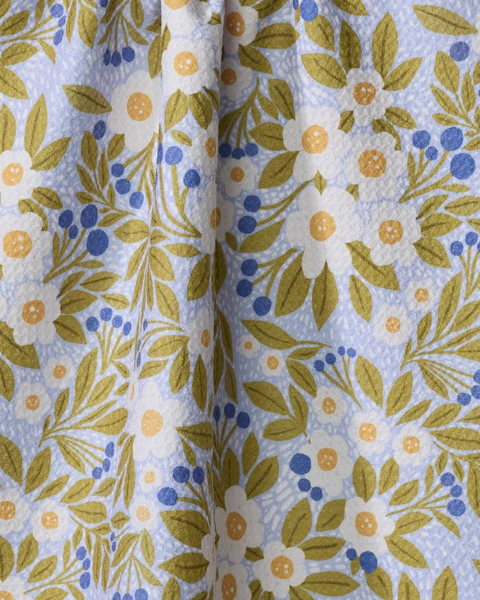 A close-up of Lisa Nelson’s Chamomile And Blue tea towel, showing green leaves, white and yellow flowers, and tiny blue berries on a light blue background. The ultra-absorbent fabric is softly gathered at the center.