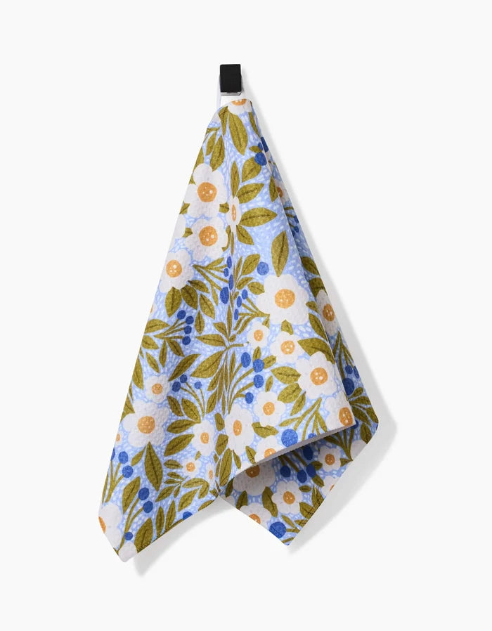 The Lisa Nelson Chamomile And Blue kitchen towel, featuring blue, green, and orange floral and leaf patterns on Geoweave fabric, hangs from a black loop against a plain white background.