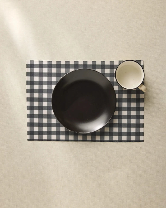 A black bowl and white mug rest on the Charcoal Gingham placemat by Geometry, neatly arranged atop a light-colored tablecloth.