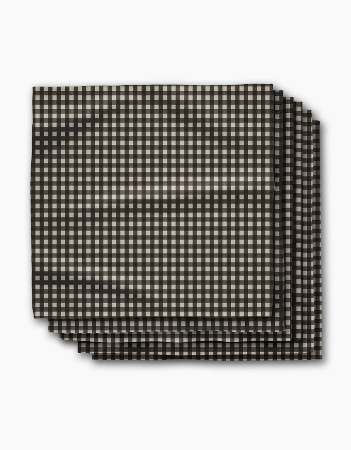 Four neatly stacked rectangular Charcoal Gingham napkins by Geometry, featuring a small black and beige check pattern, photographed on white. This eco-friendly Dinner Napkin Set is crafted from recycled materials for sustainable dining.