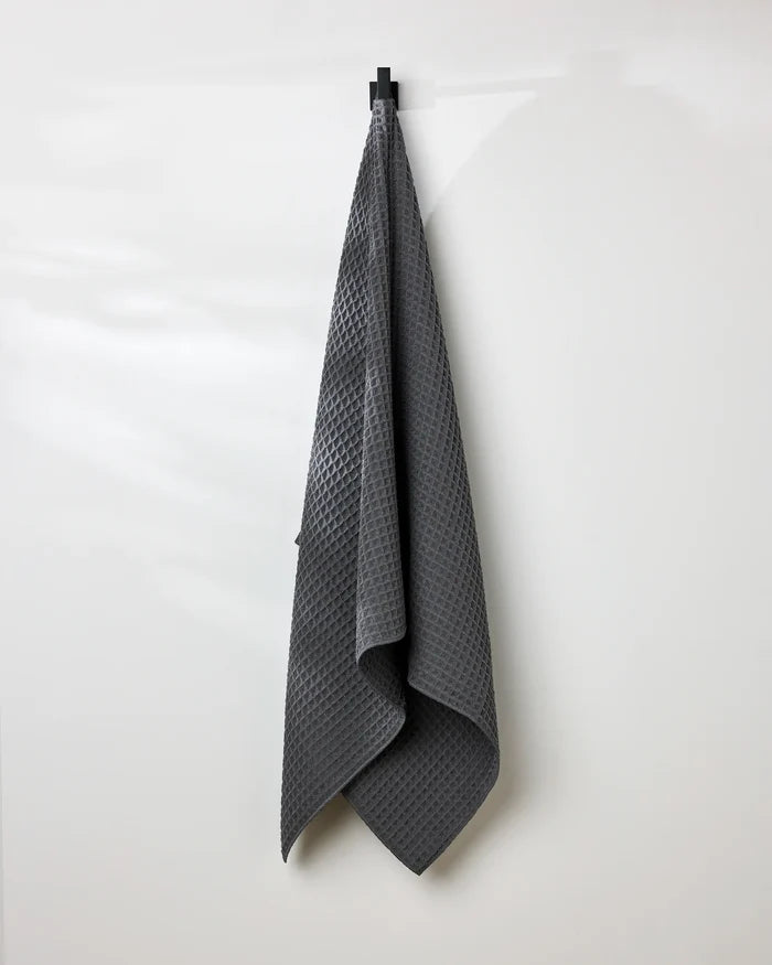 A GEOMETRY Charcoal waffle bath towel hangs neatly on a black hook against a plain white wall.