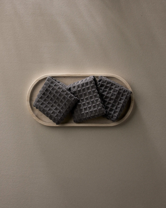 A wooden tray displays three GEOMETRY Charcoal waffle washcloths, neatly arranged on a light beige surface, with soft natural light creating gentle shadows.
