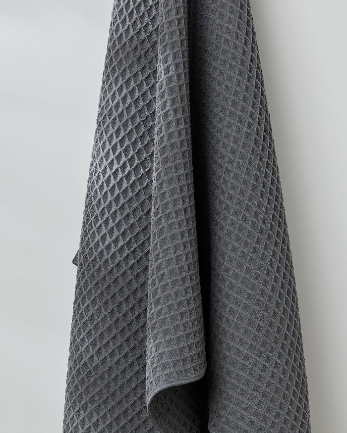 The GEOMETRY Charcoal luxury bath towel, with its soft diamond-patterned waffle texture, hangs elegantly against a light-colored wall.