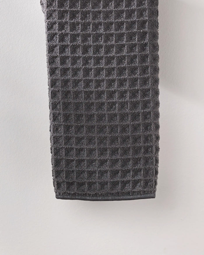 The GEOMETRY Charcoal hand towel, featuring a rich waffle texture in dark gray, hangs against a light wall to bring a touch of luxury to your space.