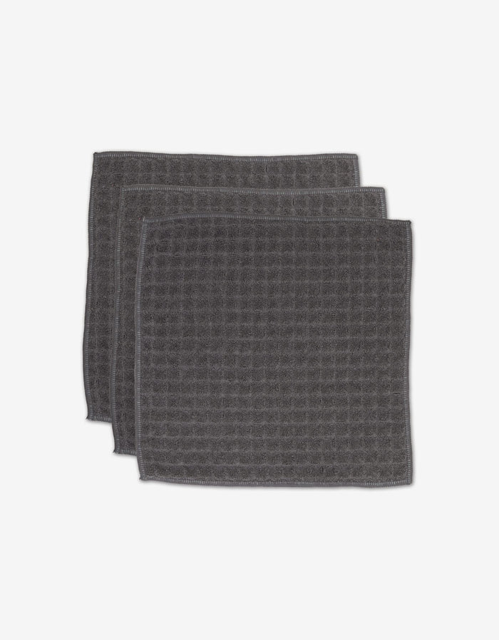 Three GEOMETRY Charcoal washcloths, featuring a dark gray color and square waffle design, are stacked with slight overlap on a white background.