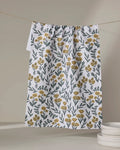 The Charolette Yellow tea towel by Charlie Rowan Designs, featuring a yellow and green floral pattern, hangs on a clothesline near a stack of white plates against a neutral background.