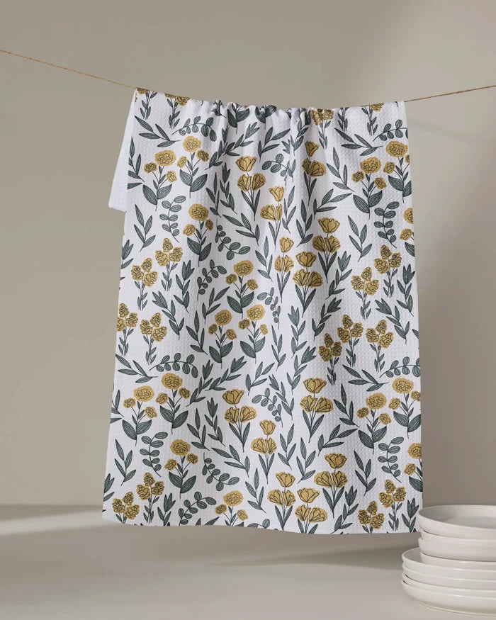 The Charolette Yellow tea towel by Charlie Rowan Designs, featuring a yellow and green floral pattern, hangs on a clothesline near a stack of white plates against a neutral background.