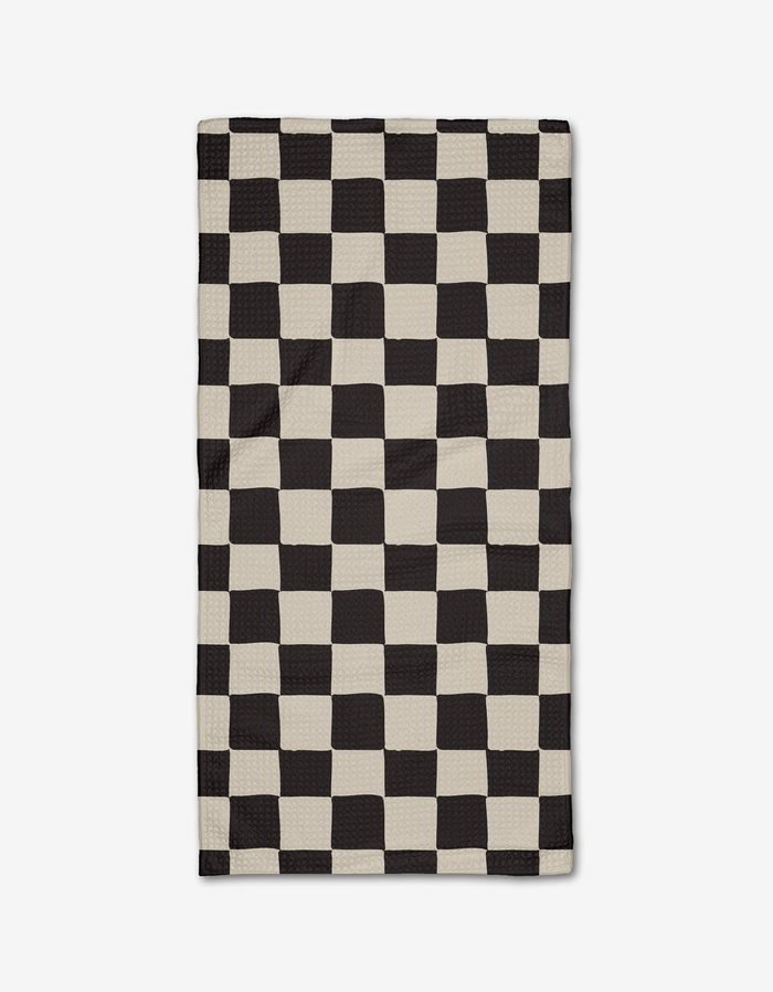 The Checkmate Bar Towel by Danelys Sidron, made of ultra-absorbent Geoweave fabric, showcases a black and off-white wavy checkerboard design that creates an optical illusion and is pictured flat on a white background.