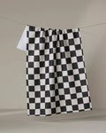 The Checkmate kitchen towel by Danelys Sidron features a classic black and white checkerboard pattern and hangs against a light background. Crafted from recycled materials, it combines style and sustainability.