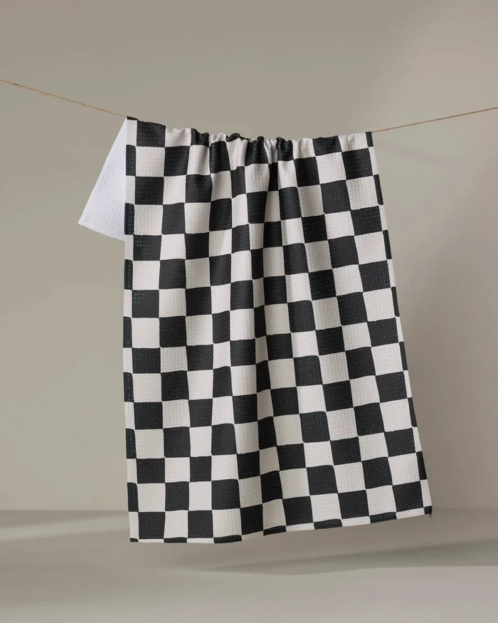 The Checkmate kitchen towel by Danelys Sidron features a classic black and white checkerboard pattern and hangs against a light background. Crafted from recycled materials, it combines style and sustainability.
