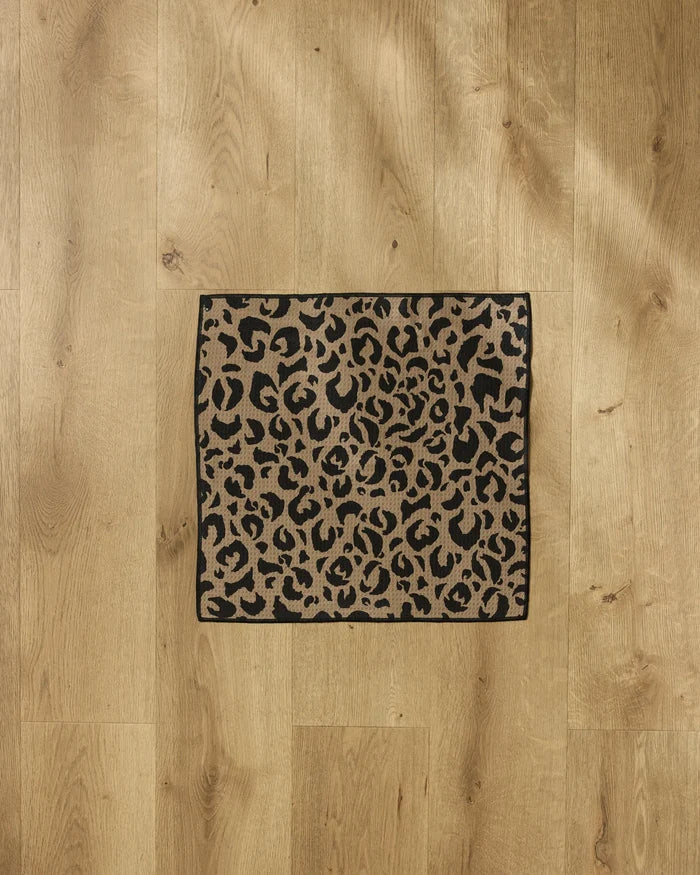 A small rectangular Cheetah Hound Paw Towel by Geometry, featuring a black and brown leopard print, is placed on a light wooden floor.