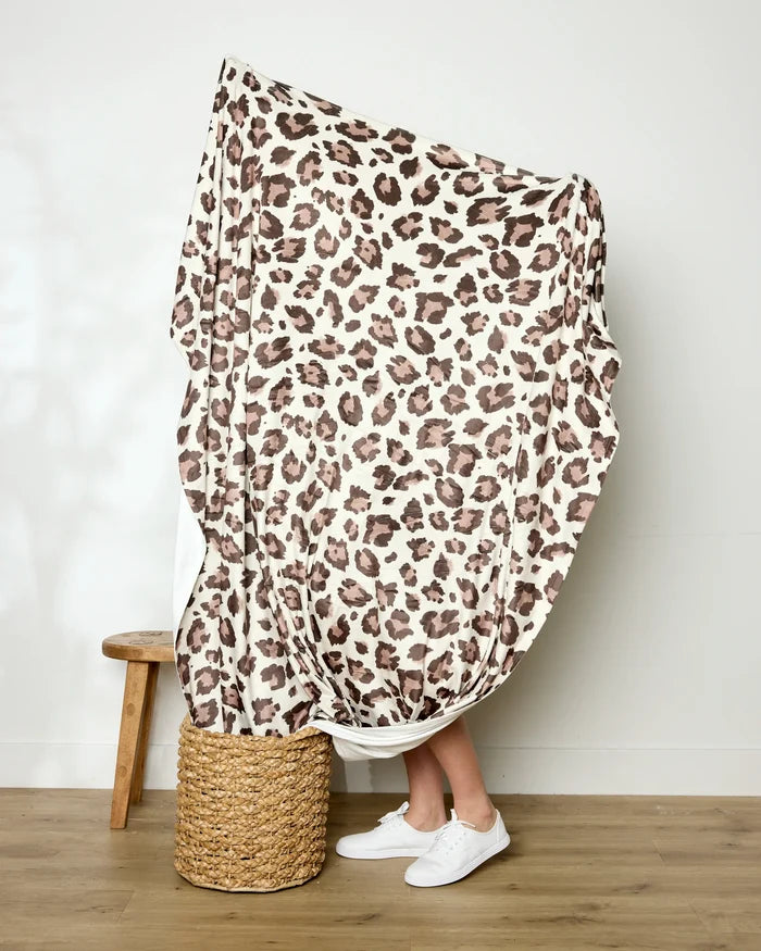 A person stands on a woven basket mostly covered by Geometry's Cheetah throw blanket, showcasing its bold leopard print. Only their lower legs and white sneakers are visible. A wooden stool is nearby on the wooden floor against a white wall.