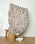 A person stands on a woven basket mostly covered by Geometry's Cheetah throw blanket, showcasing its bold leopard print. Only their lower legs and white sneakers are visible. A wooden stool is nearby on the wooden floor against a white wall.