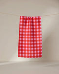 The GEOMETRY Cherry Gingham ultra-absorbent towel in red and pink checks hangs neatly on a clothesline against a plain, light background.