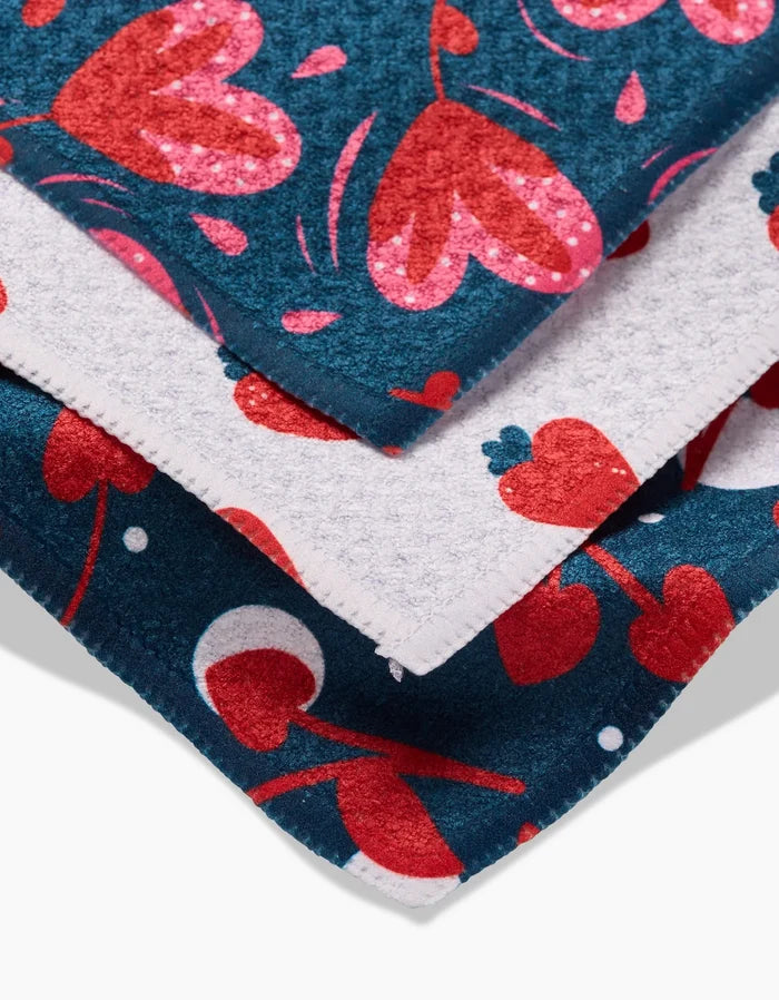 Close-up of two Cherry Love dishcloths by Charlie Rowan Designs, crafted from soft Geoweave fabric with vibrant heart and cherry patterns in red, pink, and white on blue. Zigzag-cut edges add a playful touch to the textured cloths.