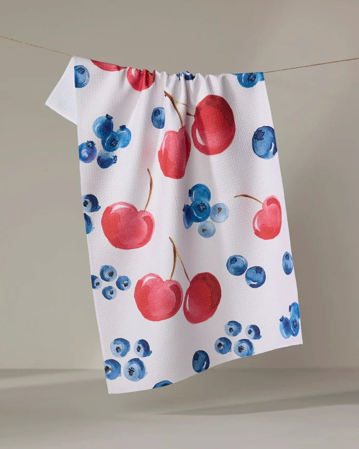 The "Cherry On Top" kitchen towel by Elena Fay, made from white recycled fabric and featuring red cherries and blue blueberries, hangs on a clothesline against a neutral background.