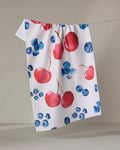 The "Cherry On Top" kitchen towel by Elena Fay, made from white recycled fabric and featuring red cherries and blue blueberries, hangs on a clothesline against a neutral background.