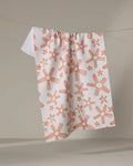 The Chunky Striped Flower tea towel by Coit Creative, made from recycled materials, features an orange floral pattern and hangs on a thin clothesline against a neutral background.