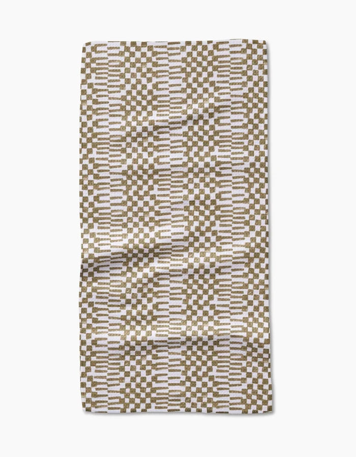 The Studio Jeannot Chutes Avocado is a rectangular bar towel made from Geoweave fabric, featuring a geometric pattern of brown and white rectangles with horizontal white lines on a neutral background. It is shown slightly wrinkled on a white surface.