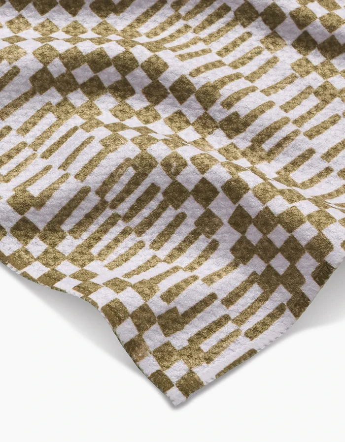A close-up of Studio Jeannot’s Chutes Avocado bar towel showcases its Geoweave fabric in an olive green and white geometric pattern of alternating squares and elongated shapes, with a gently draped, textured appearance.