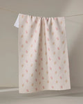 The Cindy tea towel by Kelsi Fullmer features a cream color, subtle textured pattern, and small pink flower prints. Made from recycled materials, it hangs neatly over a clothesline against a neutral background.