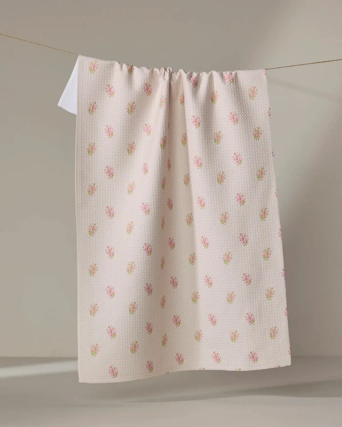 The Cindy tea towel by Kelsi Fullmer features a cream color, subtle textured pattern, and small pink flower prints. Made from recycled materials, it hangs neatly over a clothesline against a neutral background.