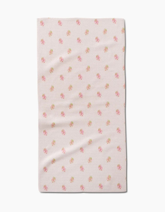The Cindy bar towel by Kelsi Fullmer features light pink fabric with a subtle textured pattern and scattered pink and yellow floral prints. Made from recycled materials, the towel is shown flat on a white background with gentle folds.