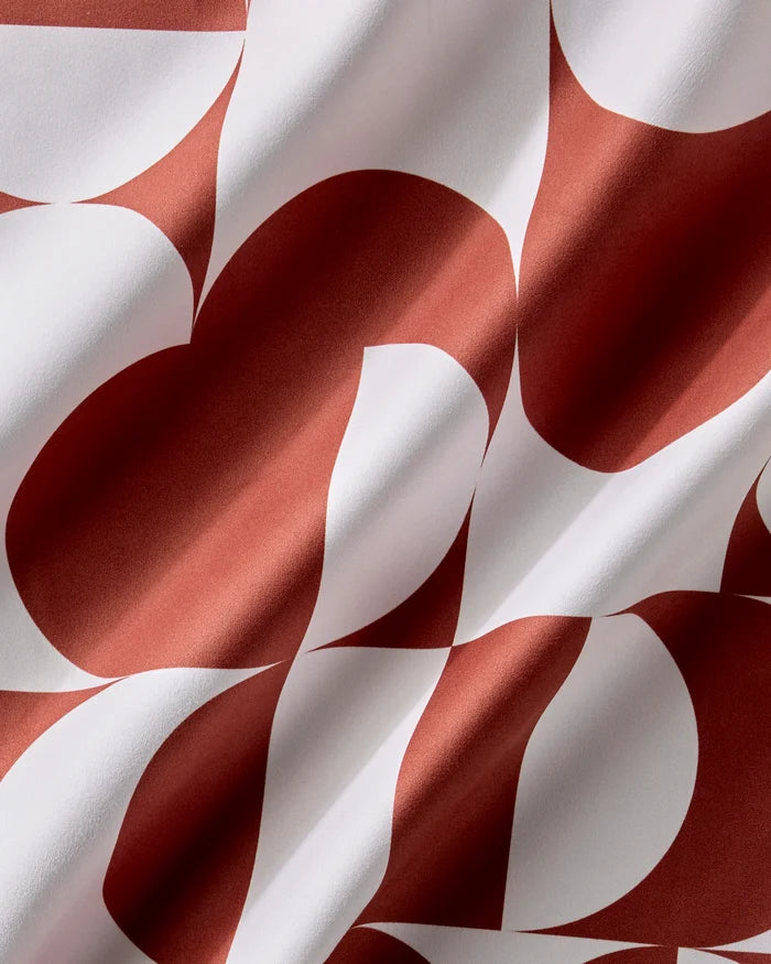 A close-up of Geometry's Circle Playground reusable napkins in rust red and white shows curved and angular patterns. The gently folded fabric creates dynamic shadows and highlights across the bold geometric design.