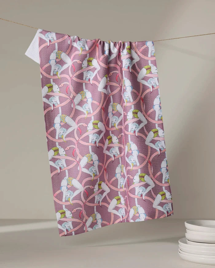 The Circus kitchen towel by Maayan Alper-Swan, featuring eco-friendly fabric and a playful print of women in white leotards and yellow hats with pink ribbons, hangs on a line near stacked white bowls.
