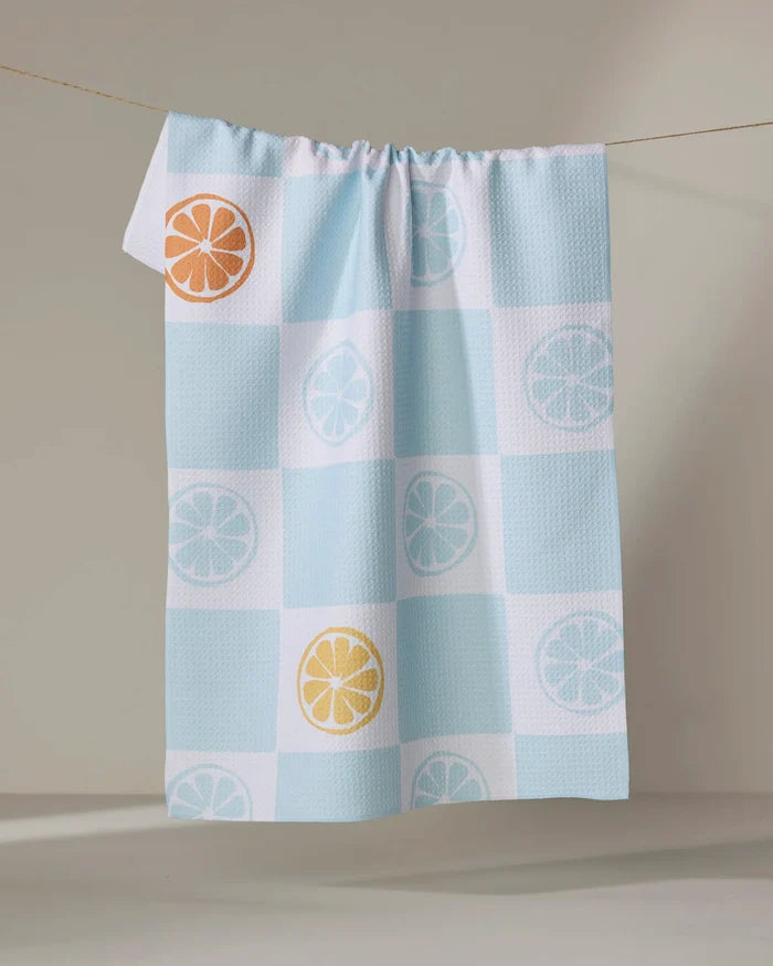 The Citrus Blue tea towel by Janna Sue Design hangs on a clothesline, showcasing blue and white checks with orange, yellow, and light blue citrus slice accents on some squares.