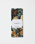 A folded eco-friendly Geometry Kitchen Tea Towel in the Classic Floral dark print with yellow, green, and rust accents, wrapped in a white label displaying the brand name. Set against a white background.