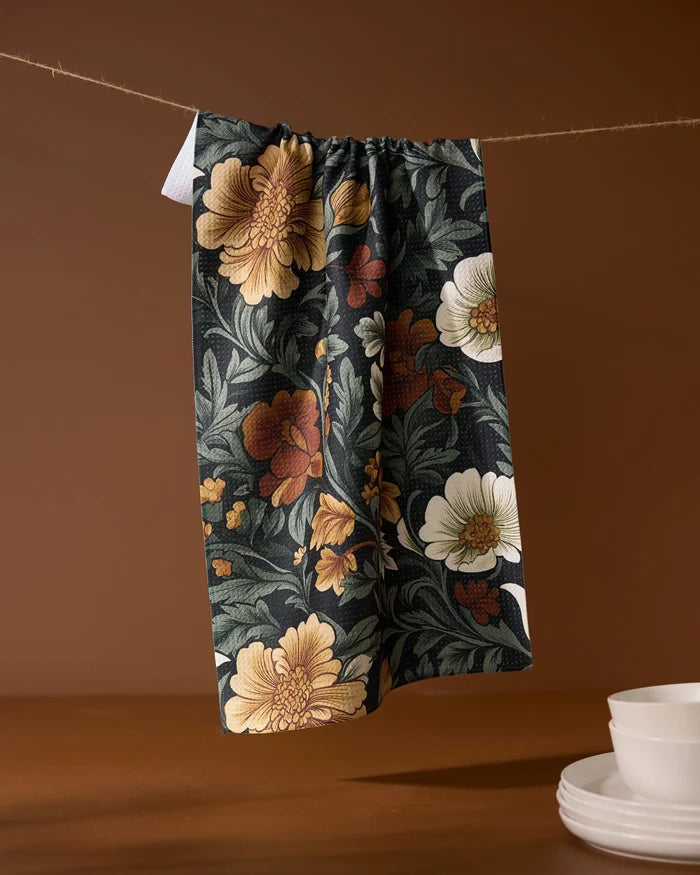 The Geometry Classic Floral eco-friendly kitchen towel, featuring yellow, white, and red flowers, hangs on a string against a brown backdrop with stacked white bowls and plates on the right.