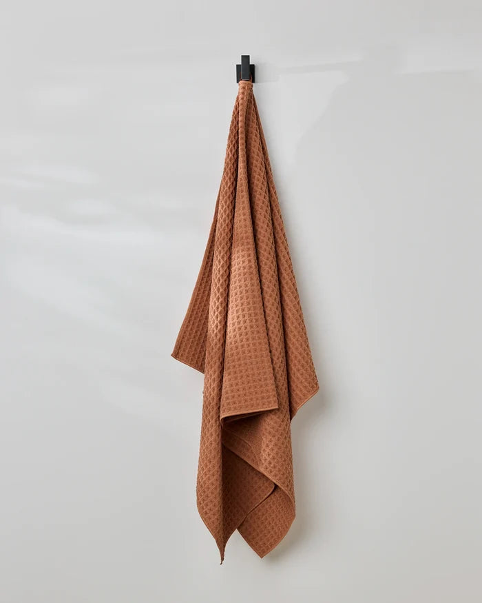 A GEOMETRY Clay waffle bath towel hangs on a black hook against a plain, light gray wall.