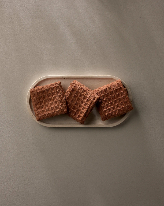 Three square chocolate wafers from GEOMETRY’s Clay collection are neatly arranged on a light oval wooden tray, their waffle-like pattern shown against a plain taupe background.