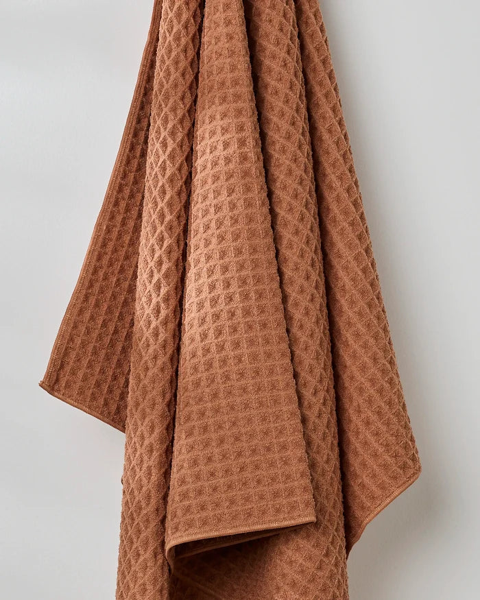 The GEOMETRY Clay bath towel, featuring a brown waffle texture, hangs neatly against a plain white wall.