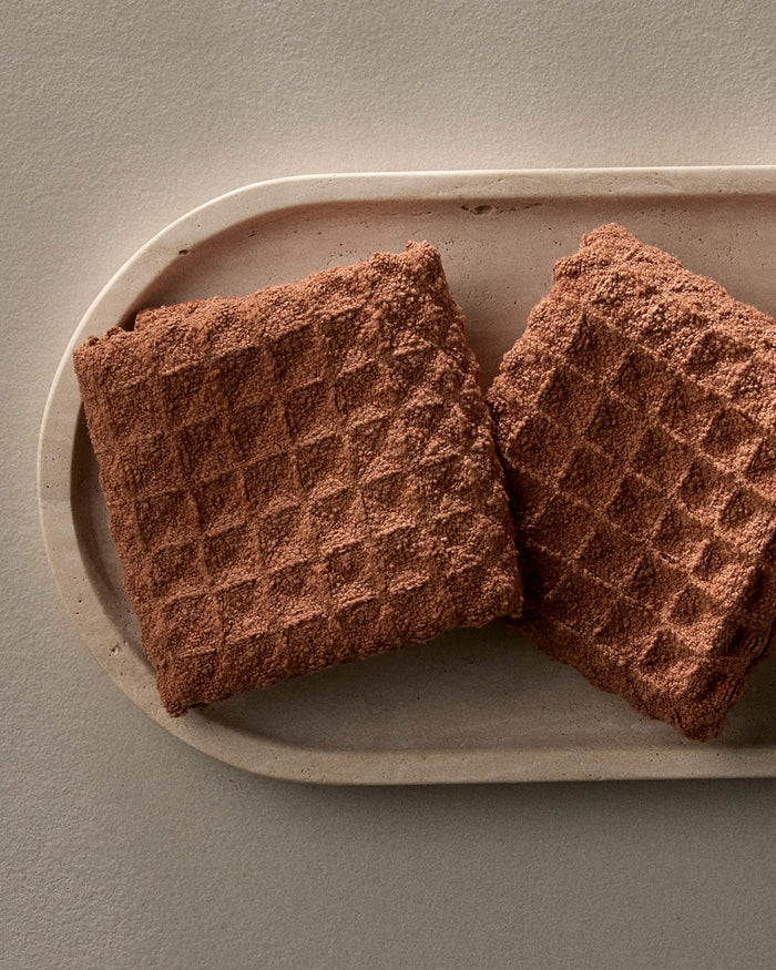 Two GEOMETRY Clay chocolate wafer biscuits, featuring a grid-like surface, are placed side by side on an oval beige ceramic plate against a neutral background, their textured tops resembling a waffle-patterned washcloth.