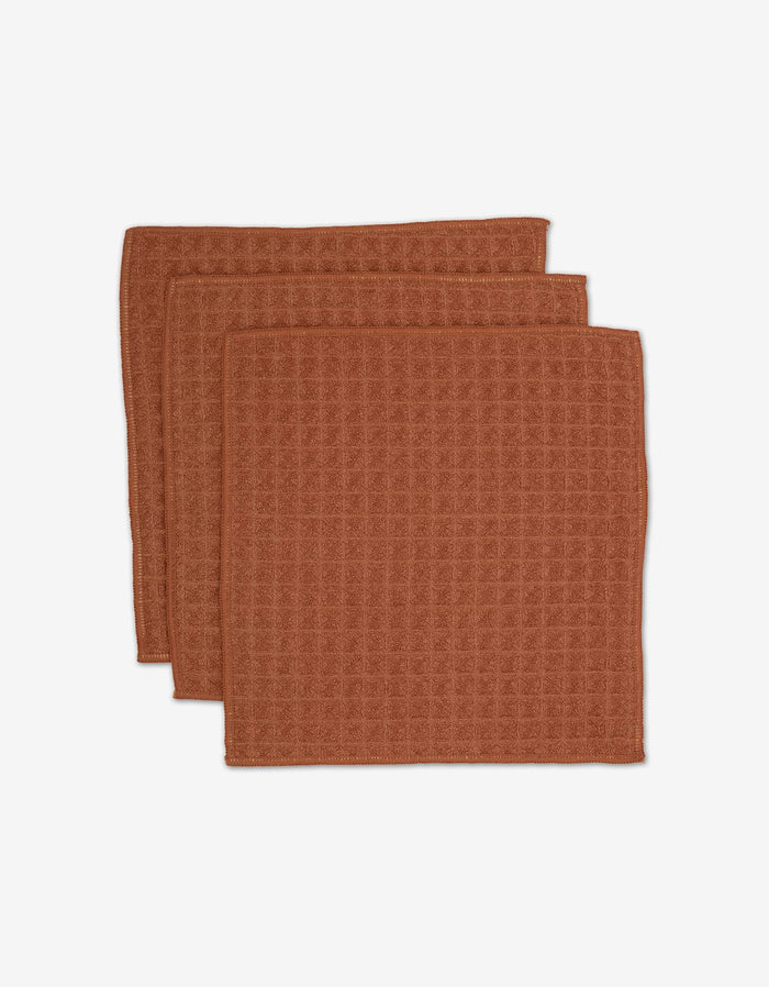 Three folded brown waffle-textured absorbent washcloths, stacked and slightly overlapping on a white background. GEOMETRY’s Clay washcloths from the Waffle Bath Collection.