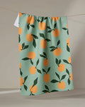 Purely Kaylie’s “Clementine Sky” tea towel, made from recycled materials, features orange fruit and dark green leaf patterns on light green fabric and hangs against a neutral background.