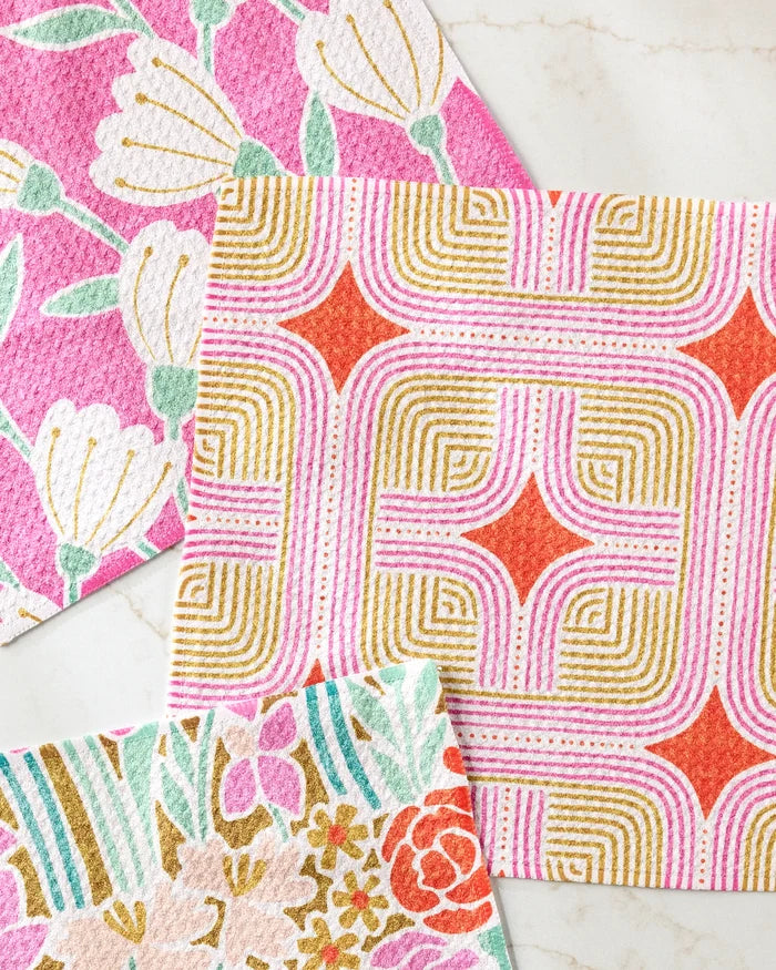 Three colorful Clemmie Donna and Janae dishcloths by Amy MacCready are displayed—one with pink and white flowers, one with geometric orange, pink, and gold shapes, and another featuring pastel floral patterns.