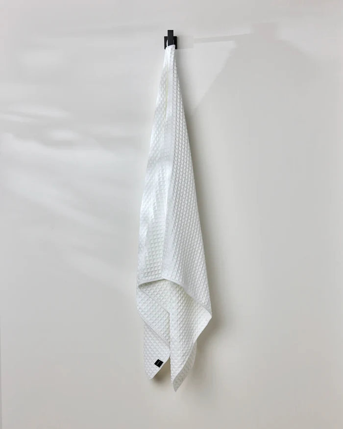A GEOMETRY Cloud white waffle bath towel hangs neatly from a black hook on a plain, light-colored wall.