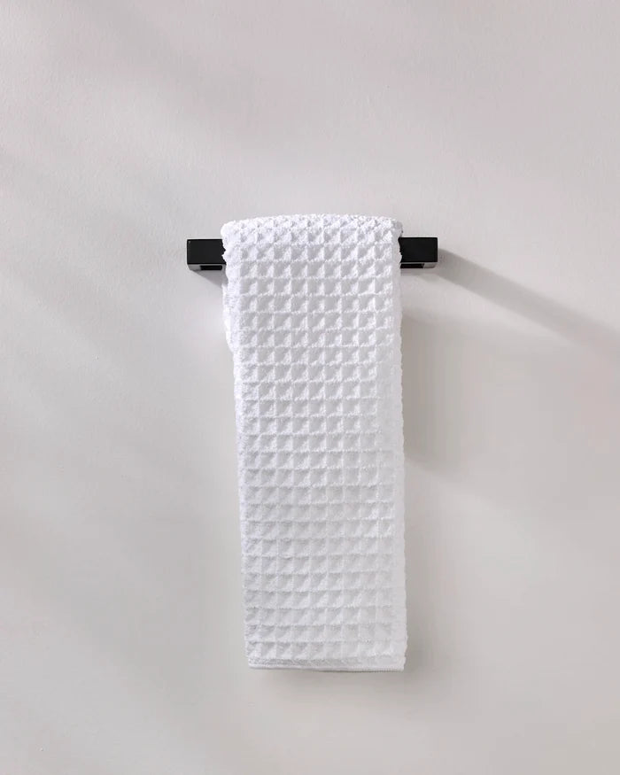 The GEOMETRY Cloud hand towel, made from recycled materials, in white waffle texture, hangs neatly on a black towel bar fixed to a light gray wall.
