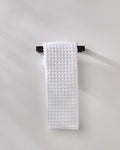 The GEOMETRY Cloud hand towel, made from recycled materials, in white waffle texture, hangs neatly on a black towel bar fixed to a light gray wall.
