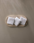 Three "Cloud" waffle washcloths by GEOMETRY, in white and featuring a textured grid pattern, are arranged on a light wooden oval tray against a plain beige background.