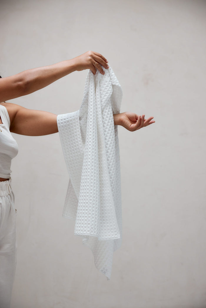 A person dressed in white holds up the GEOMETRY Cloud Waffle Bath Towel with both hands against a plain light background, showing only their upper body and arms.