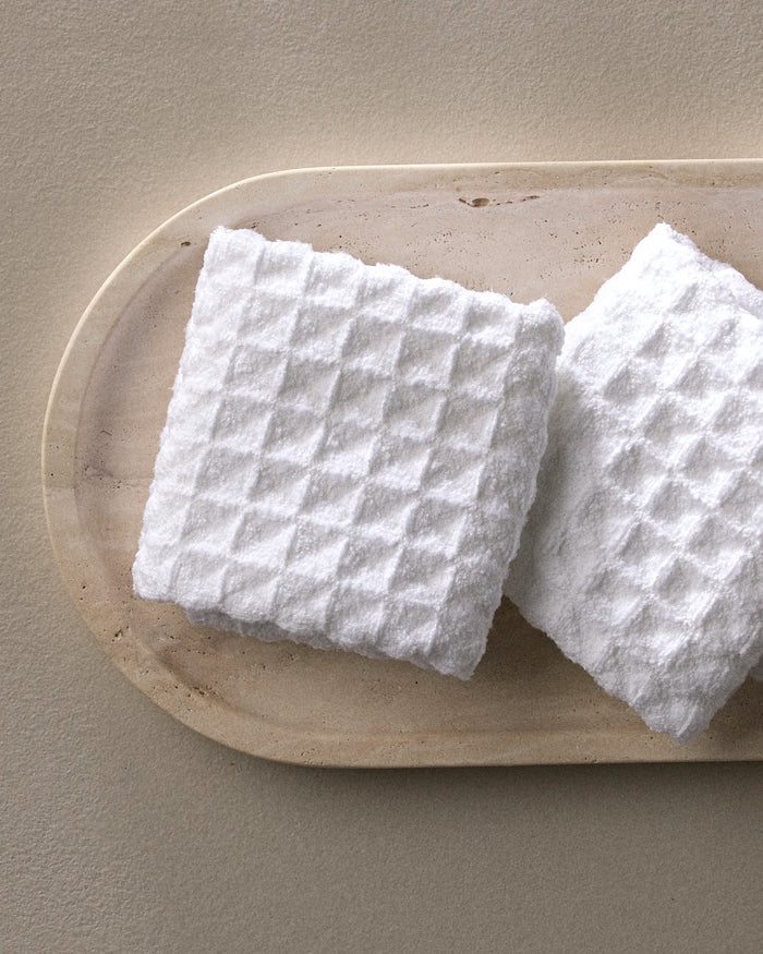 Two white, textured Cloud washcloths by GEOMETRY rest on an oval, light marble tray atop a beige surface. Crafted from recycled materials, they bring eco-conscious luxury and understated elegance to your space.